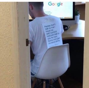 Guy working at a computer with a sign on his back that says, "Please don't talk to me I have no self-control and will talk to you for two hours and get no work done."